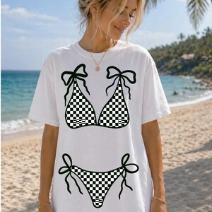May include: White t-shirt with a black and white chequered bikini design. The bikini top and bottom are outlined with black bows. A casual, beach-themed apparel item.