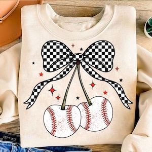 May include: A cream-colored sweatshirt with a black and white checkered bow and two red cherries with baseball stitching. The cherries have stems with green leaves and are surrounded by red stars and white sparkles.