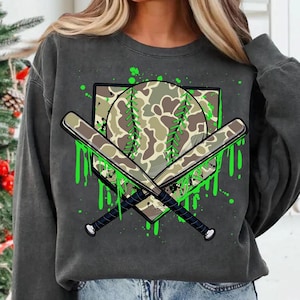May include: A grey sweatshirt with a green, white and brown camouflage baseball graphic. The graphic features a baseball, two crossed bats and a home plate with green paint splatters.