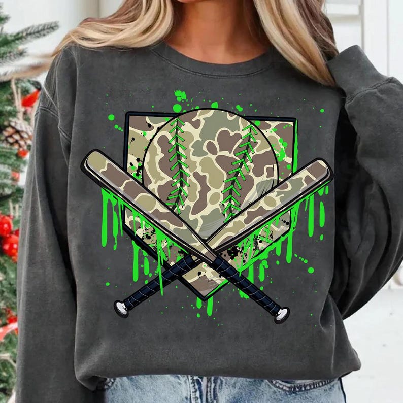 May include: A grey sweatshirt with a green, white and brown camouflage baseball graphic. The graphic features a baseball, two crossed bats and a home plate with green paint splatters.