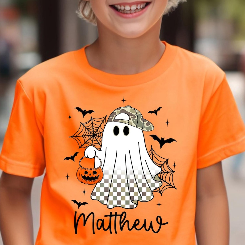 Puede incluir: Camiseta naranja brillante con un fantasma de dibujos animados que lleva una gorra de camuflaje y una calabaza. El dise&ntilde;o incluye murci&eacute;lagos, telara&ntilde;as y el nombre "Matthew". Una camiseta gr&aacute;fica con tem&aacute;tica de Halloween.