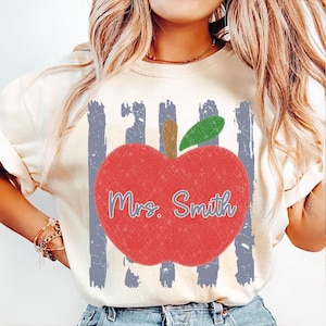 Puede incluir: Camiseta color crema con un gran gráfico de manzana roja con tallo marrón y hoja verde. La manzana tiene el texto "Mrs. Smith" en escritura azul. Rayas azules verticales detrás de la manzana. La camiseta es de estilo casual.