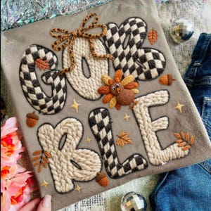 May include: A grey fabric featuring the embroidered words "GIVE & BLESSED". The letters are a mix of checkered and knitted textures. The design includes a turkey, acorns, leaves, and stars. A bow is tied at the top.