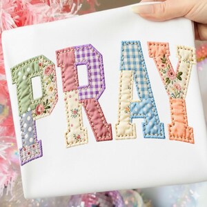 May include: White fabric with the word "PRAY" appliqued in patchwork letters. Each letter features a different floral or patterned fabric, including gingham, polka dots, and floral prints. The letters are outlined with a contrasting stitch.