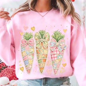 Op de afbeelding: Roze sweatshirt met een wortelontwerp. De wortels hebben patchworkpatronen, groene toppen en decoratieve strikken. Gouden harten en spikkels dragen bij aan het lente-thema.