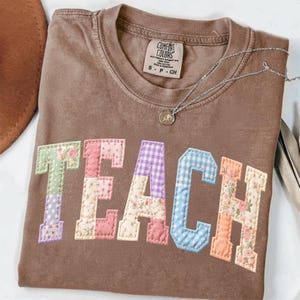 Op de afbeelding: Een bruin Comfort Colors T-shirt met het woord "TEACH" als applicatie op de voorkant van patchwork stof. De letters hebben verschillende patronen, waaronder bloemen, ruitjes en stippen. Een zilveren ketting met een kleine hanger is ook zichtbaar.