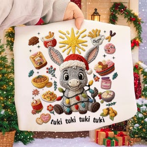 May include: A white pillow featuring a gray donkey wearing a Santa hat and string lights, surrounded by embroidered food items. The text "tuki tuki tuki tuki" is at the bottom. The pillow is held in front of a decorated house.