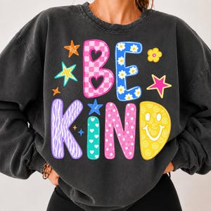 Op de afbeelding: Een antracietgrijze sweater met ronde hals en de tekst BE KIND in grote, veelkleurige letters. De letters hebben diverse patronen, waaronder roze ruitjes, blauwe madeliefjes, paarse zebrastrepen, turquoise hartjes, roze stippen en een gele smiley.