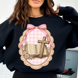 May include: Black sweatshirt featuring a pink and white gingham oval design with a pink bow, a wooden cross, and an open book. The design includes floral accents and a decorative border.