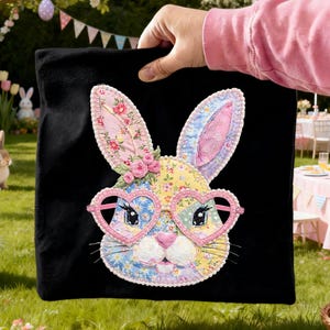 May include: Black square pillow featuring a colorful patchwork bunny face with heart-shaped glasses. The bunny has floral patterned ears and a yellow, blue, and pink face. Pink roses adorn the bunny's head, creating a whimsical Easter theme.