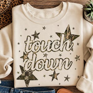 May include: A cream-colored crewneck sweatshirt featuring the text "touch down" in a white font with a dark outline. The design includes multiple stars of varying sizes with a camouflage pattern, scattered across the front of the garment.