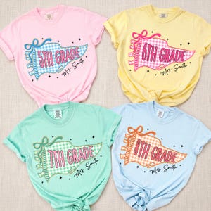 May include: Four short-sleeve t-shirts in pastel pink, yellow, mint green, and light blue. Each features a graphic of a gingham pennant flag with the text 5th Grade, 6th Grade, 7th Grade, or 8th Grade, plus the handwritten name Mrs. Smith.