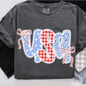 Puede incluir: Sudadera gris oscuro con la palabra "USA" en un diseño patriótico. Las letras están decoradas con estrellas azules, cuadros rojos y patrones florales azules, delineados con encaje blanco y lazos rojos.