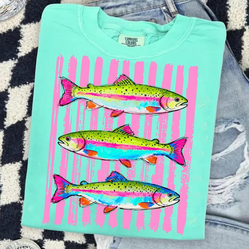 May include: A light teal Comfort Colors sweatshirt featuring three colorful fish illustrations. The fish have green bodies, pink fins, and rainbow stripes. Vertical pink brushstrokes are behind the fish. The shirt is folded on a denim surface.