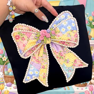 May include: Black top featuring a large, colorful bow applique. The bow is made of patchwork fabric with floral and gingham patterns in yellow, pink, and blue. The bow is outlined with white pearls and adorned with small pink roses.