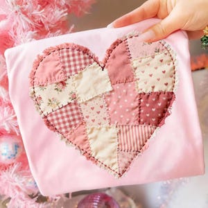 Pode incluir: Uma camisa rosa claro com um design de coração de patchwork. O coração é composto por vários tecidos rosa e creme com padrões como xadrez, florais e bolinhas. O coração é contornado com uma borda desfiada.