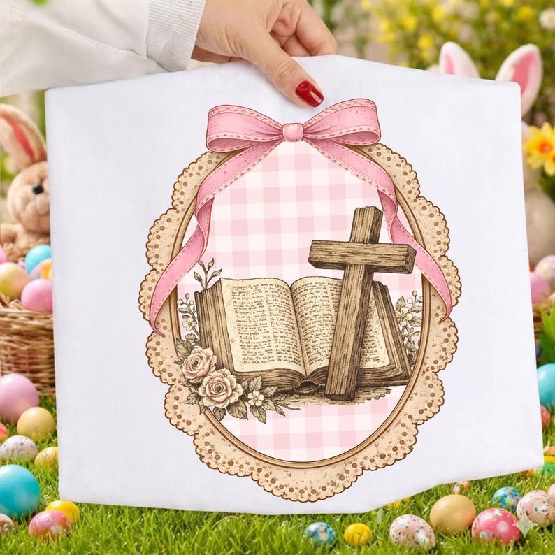 May include: White fabric with a pink gingham background, a wooden cross, and an open book illustration. A pink ribbon and a decorative frame with floral accents surround the image. The design is held by a person.