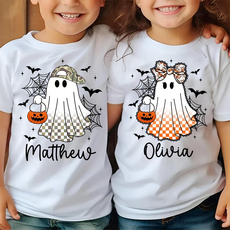 Puede incluir: Camisetas blancas con dise&ntilde;os de fantasmas de Halloween. Un fantasma lleva una gorra de camuflaje, el otro un lazo. Ambos sostienen bolsas de golosinas de calabaza naranja. Murci&eacute;lagos negros y telara&ntilde;as rodean a los fantasmas. Los nombres "Matthew" y "Olivia" est&aacute;n impresos debajo.