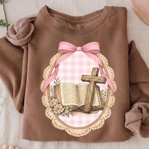 May include: A brown sweatshirt featuring a detailed graphic. The graphic includes an open book, a wooden cross, and a pink bow. The design is set within a decorative oval frame with a pink gingham background and floral accents.
