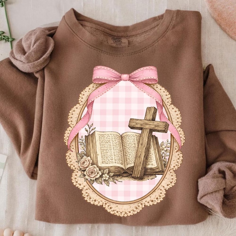 May include: A brown sweatshirt featuring a detailed graphic. The graphic includes an open book, a wooden cross, and a pink bow. The design is set within a decorative oval frame with a pink gingham background and floral accents.