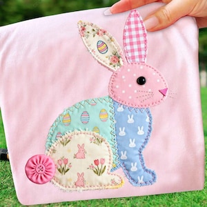 Könnte beinhalten: Ein rosa Stoff-Osterhasen-Applikation mit Patchwork-Details. Der Hase hat ein rosa gepunktetes Gesicht, ein kariertes Ohr und ein Blumenohr. Der Körper hat Abschnitte mit Ostereiern, Kaninchensilhouetten und floralen Mustern. Unten links befindet sich ein rosa Stoffknopf.