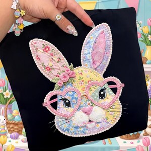 May include: Black tote bag featuring a colorful patchwork bunny face with heart-shaped glasses and floral accents. The bunny has pink, yellow, and blue floral patterns. The bag is perfect for carrying books, groceries, or other items.