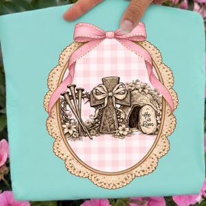 May include: A light blue Easter design with a pink bow, cross, nails, and a tomb with the text "He is Risen." The design is set within an oval frame with a pink gingham background and a decorative border.