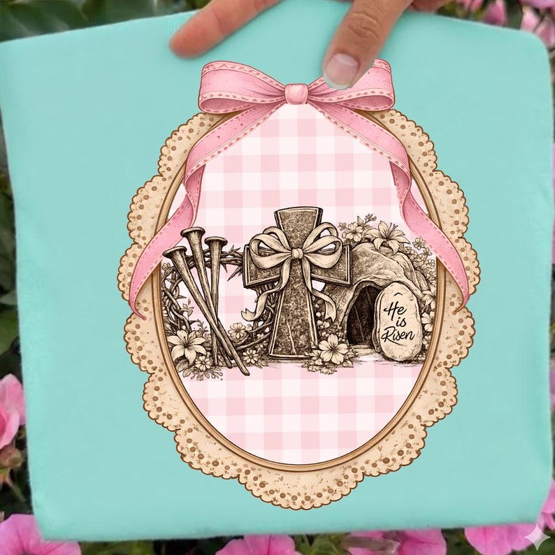 May include: A light blue Easter design with a pink bow, cross, nails, and a tomb with the text "He is Risen." The design is set within an oval frame with a pink gingham background and a decorative border.