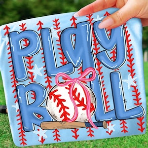 May include: A light blue towel featuring the words "PLAY BALL" in large, outlined blue letters. A baseball with a pink bow and a wooden bat are depicted. Red stitching and star accents complete the design.