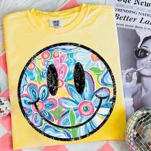 May include: Yellow Comfort Colors t-shirt featuring a large smiley face design. The smiley face is filled with a floral pattern in shades of blue, pink, and yellow, outlined in black. The shirt is folded and displayed on a pink and white checkered surface.
