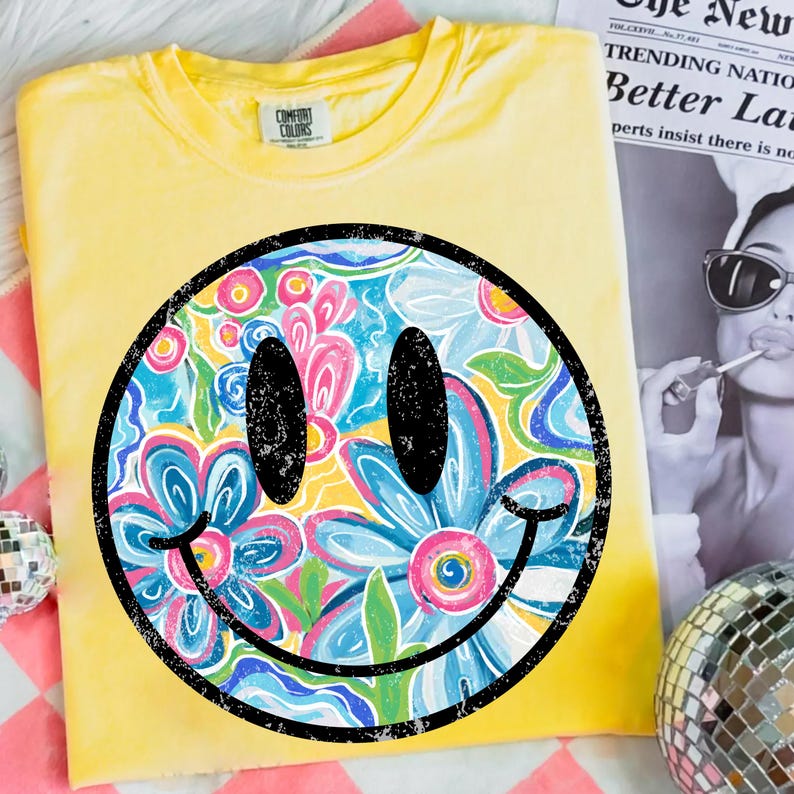 May include: Yellow Comfort Colors t-shirt featuring a large smiley face design. The smiley face is filled with a floral pattern in shades of blue, pink, and yellow, outlined in black. The shirt is folded and displayed on a pink and white checkered surface.