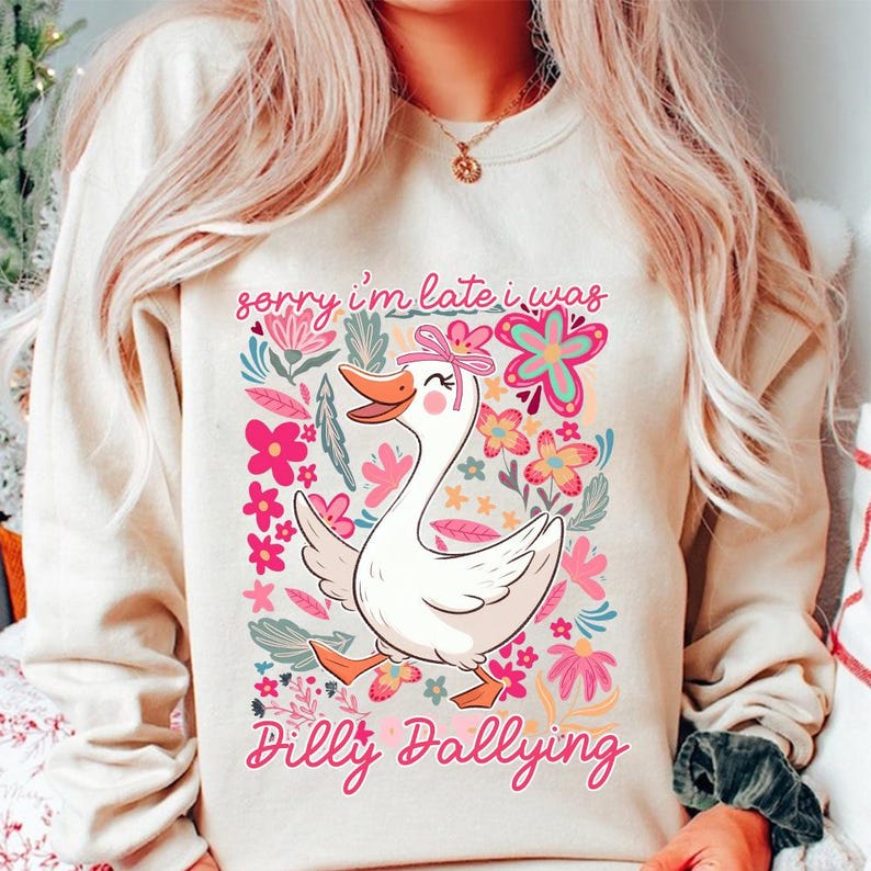 K&ouml;nnte beinhalten: Cremefarbenes Sweatshirt mit einer Cartoon-Gans mit rosa Schleife, umgeben von floralen Designs in Rosa, Blau und Gr&uuml;n. Der Text "sorry i'm late i was Dilly Dallying" ist in rosa Schrift gedruckt, was ein skurriles und verspieltes Design ergibt.