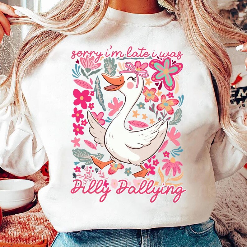 K&ouml;nnte beinhalten: Wei&szlig;es Sweatshirt mit einem Cartoon-Gans-Design, einer rosa Schleife und floralen Elementen. Der Text lautet "sorry i'm late i was dilly dallying" in rosa Schrift. Ein verspieltes und skurriles Design.