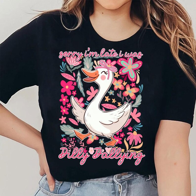 K&ouml;nnte beinhalten: Schwarzes T-Shirt mit einem Cartoon-Gans-Design, die eine rosa Schleife tr&auml;gt und von Blumen umgeben ist. Der Text "sorry i'm late i was Dilly Dallying" steht &uuml;ber und unter der Gans. Die Grafik ist auf die Vorderseite des T-Shirts gedruckt.