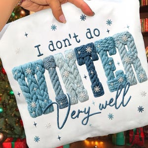 以下が含まれることがあります： 「I don't do WINTER very well」と刺繍された白いクッション。WINTERの文字は、雪の結晶のアクセントが付いた青いニットのデザインです。テキストの周りには、追加の雪の結晶と星が刺繍されています。