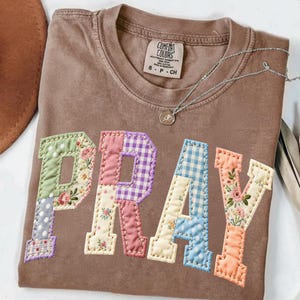 Könnte beinhalten: Ein braunes Comfort Colors T-Shirt mit dem Wort "PRAY" aus bunten Patchwork-Stoffen. Der Patchwork-Stoff umfasst Blumen-, Tupfen- und Vichy-Muster. Eine silberne Halskette mit einem kleinen Anhänger liegt auf dem Shirt.
