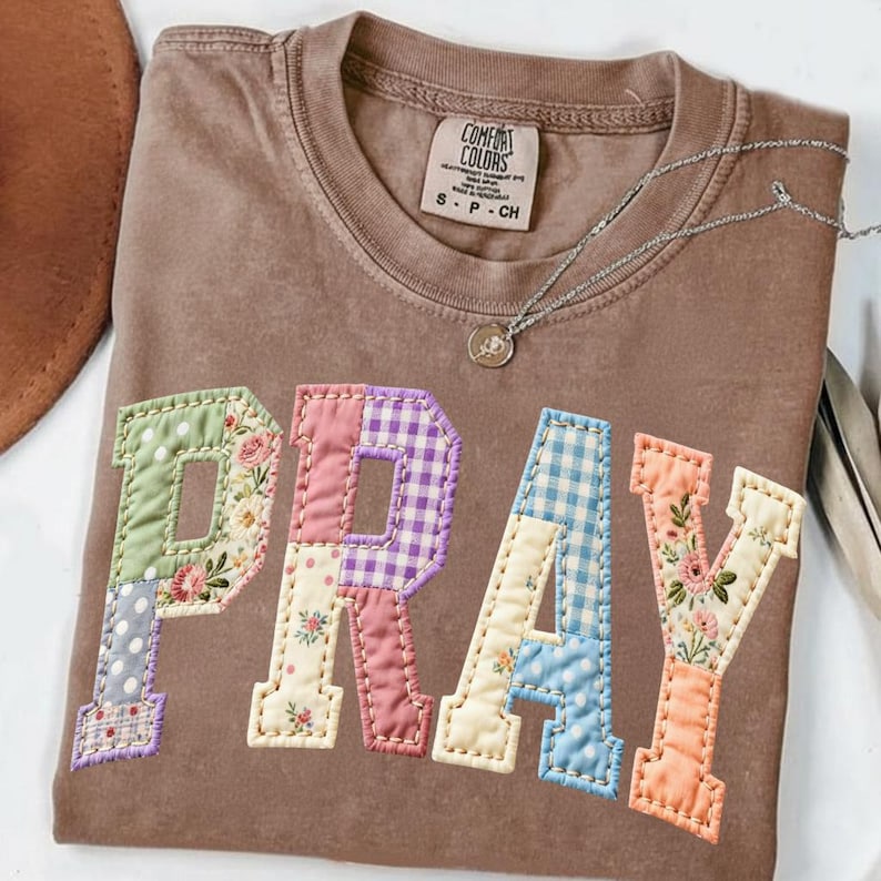 May include: A brown Comfort Colors t-shirt with the word "PRAY" appliqued in colorful patchwork fabric. The patchwork includes floral, polka dot, and gingham patterns. A silver necklace with a small pendant rests on the shirt.