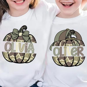 Puede incluir: Dos camisetas blancas con diseños de calabaza. Una dice "Olivia" y la otra "Oliver". Los diseños de calabaza tienen un patrón de camuflaje en tonos marrones, verdes y beige. La calabaza de Olivia tiene un lazo verde.