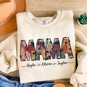 May include: Cream-colored sweatshirt with the word "MAMA" in large, floral-filled letters. Below the word are the names "Layla", "Oliver", and "Taylor". The sweatshirt is made of a soft material and is perfect for a mother.