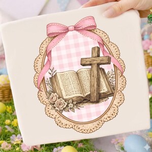 May include: A cream-colored gift box featuring a pink bow and an oval design with a cross, open book, and roses. The background of the oval is a pink and white gingham pattern. The design is framed by a scalloped lace border.