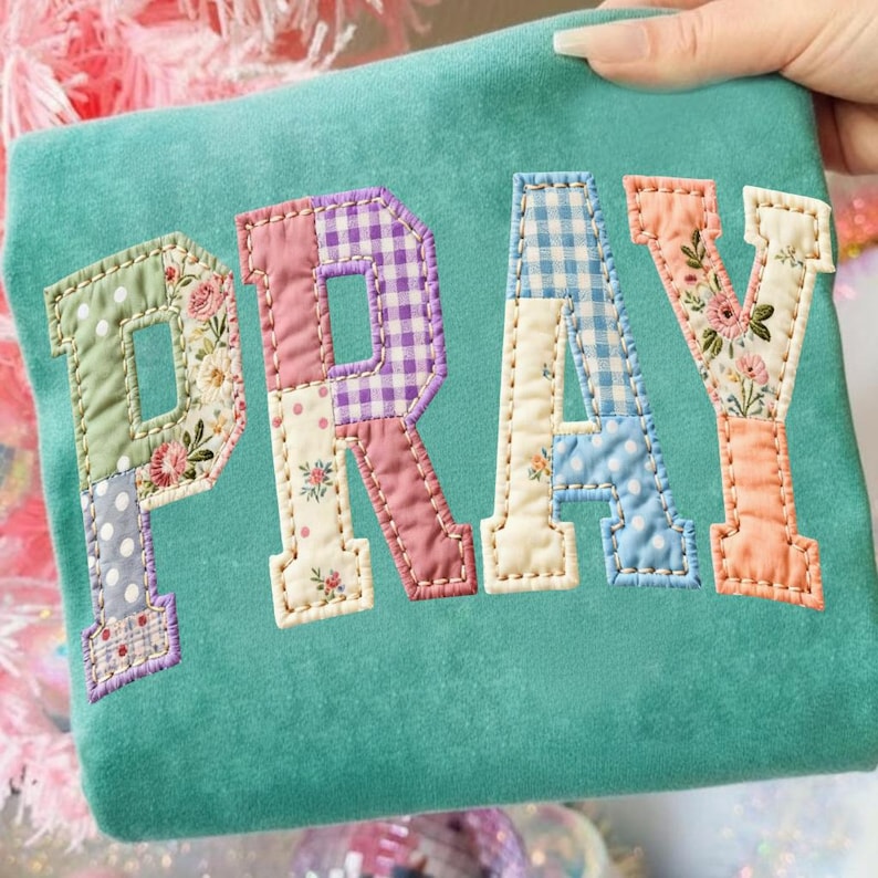 May include: A teal-colored fabric item with the word "PRAY" appliqu&eacute;d in patchwork letters. Each letter features a different floral or patterned fabric, including gingham, polka dots, and floral prints. The letters are outlined with a cream-colored stitching.