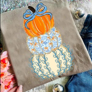 May include: A tan-colored t-shirt featuring a stacked pumpkin design. The top pumpkin is orange with a blue bow, the middle pumpkin has a blue floral pattern, and the bottom pumpkin is cream-colored with blue leaf accents.