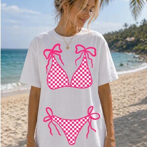 May include: White t-shirt with a pink and white chequered bikini design. The bikini top and bottom are outlined in pink with bow details. The shirt is worn on a beach with a blue sky and palm trees in the background.