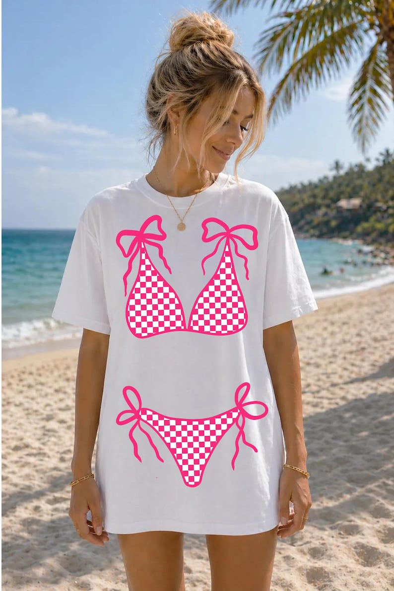 May include: White t-shirt with a pink and white chequered bikini design. The bikini top and bottom are outlined in pink with bow details. The shirt is worn on a beach with a blue sky and palm trees in the background.