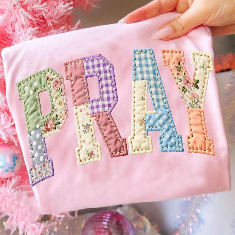 May include: A light pink shirt with the word "PRAY" appliqued on the front. The letters are made of various patterned fabrics, including floral, polka dot, and gingham, in pastel colors. The shirt is held by a hand.