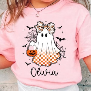 Puede incluir: Camiseta rosa con un dise&ntilde;o de fantasma de Halloween. El fantasma lleva un lazo y una cesta de calabaza. Murci&eacute;lagos negros y telara&ntilde;as rodean al fantasma. El nombre "Olivia" est&aacute; impreso en negro debajo.