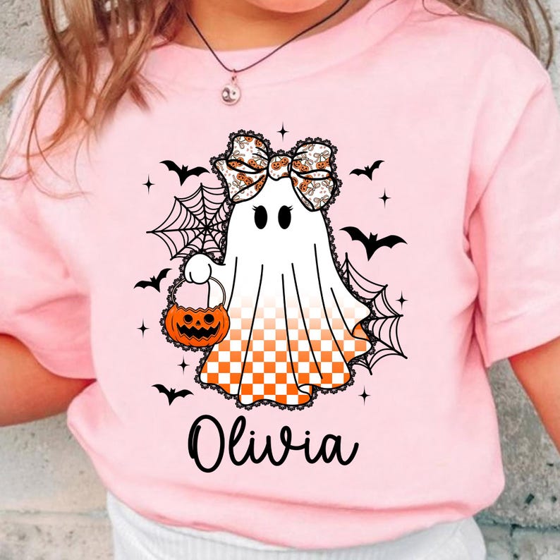 Puede incluir: Camiseta rosa con un dise&ntilde;o de fantasma de Halloween. El fantasma lleva un lazo y una cesta de calabaza. Murci&eacute;lagos negros y telara&ntilde;as rodean al fantasma. El nombre "Olivia" est&aacute; impreso en negro debajo.