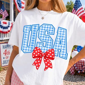 May include: White t-shirt with the word "USA" in blue gingham letters. A red bow with white polka dots is centered below the text. The shirt is likely for the 4th of July.