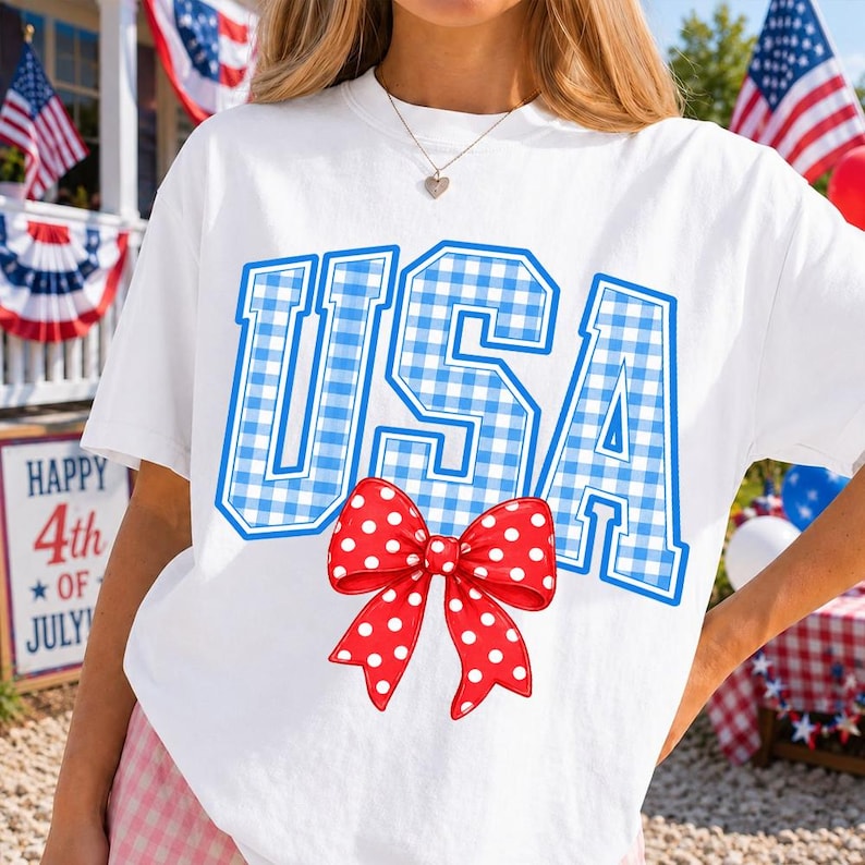 May include: White t-shirt with the word "USA" in blue gingham letters. A red bow with white polka dots is centered below the text. The shirt is likely for the 4th of July.