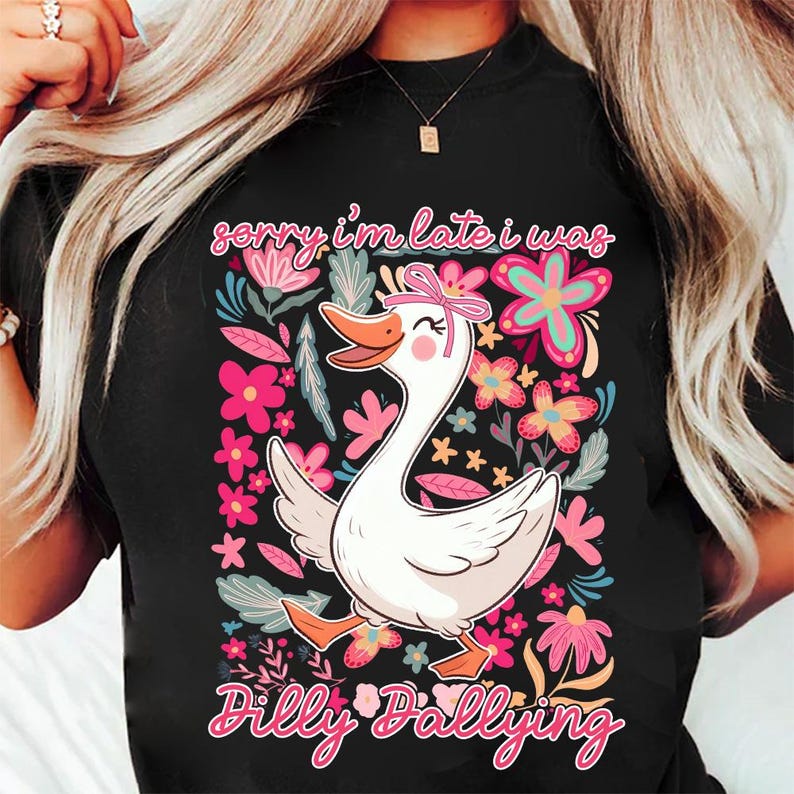 K&ouml;nnte beinhalten: Schwarzes T-Shirt mit einer skurrilen Grafik einer wei&szlig;en Gans mit rosa Schleife, umgeben von bunten Blumen. Der Text "Sorry I'm late I was dilly dallying" ist in Rosa und Wei&szlig; dargestellt.